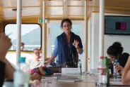Frau hält Vortrag auf einem Boot, Menschen hören zu.