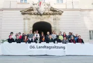 Frauen vor einem Gebäude mit Banner "Oö. Landtagssitzung", lächelnd und bunt gekleidet.