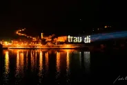 Nächtliche Stadtansicht mit beleuchtetem Schriftzug "trau di" über Wasser.