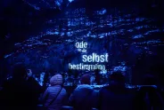 Personen vor beleuchteter Felswand mit Text "Ode an die Selbstbestimmung".