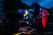 Frau mit roten Mantel bei Nacht an Filmkamera, dunkle Berglandschaft im Hintergrund.