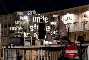 Zwei Personen auf einer Bühne, projizierte Wörter "unite" und "strike" im Hintergrund.