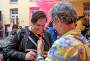 Zwei Personen, eine bindet der anderen ein Halstuch, lächelnd im bunten Innenhof.