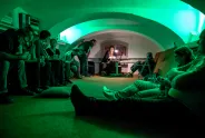 Menschen in grün beleuchtetem Raum, einige sitzen, andere stehen.
