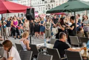 Menschenmenge auf einem Platz, einige halten Schilder, im Vordergrund Personen an Tischen.