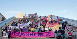 Menschenmenge hält Schilder auf Treppe, Banner mit „Feministischer Protest“.