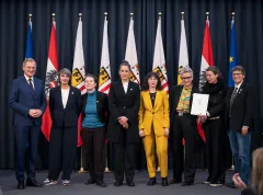 Eine Gruppe von acht Personen steht vor nationalen und regionalen Flaggen auf einer Bühne.