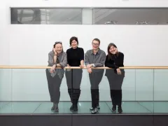 Vier Frauen lehnen lächelnd an einem Glasgeländer in einem modernen Gebäude.