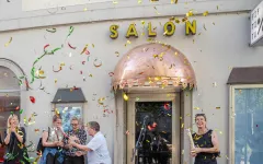 Menschen feiern vor einem Gebäude mit der Aufschrift "SALON", bunte Konfetti fliegen.