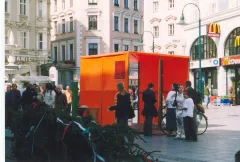 Rote Informationshütte auf belebtem Stadtplatz. Menschen stehen davor und unterhalten sich.