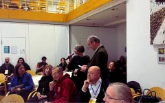 Versammlung in Konferenzraum, Person spricht in Mikrofon, Zuhörer aufmerksam.
