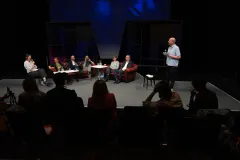 Diskussionsrunde mit sieben Personen, eine spricht stehend; Theaterbühne, abgedunkelt.