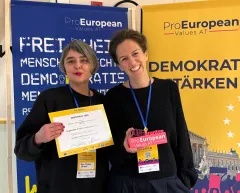 Zwei Frauen lächeln mit Zertifikat und Ausweis vor ProEuropean Plakaten.