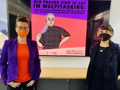 Zwei Frauen mit Masken vor feministischem Plakat zur Gender Pay Gap.