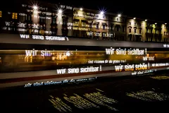 Lichter mit "Wir sind sichtbar!" auf vorbeifahrender Tram bei Nacht.