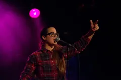 Frau mit Brille singt auf der Bühne, trägt ein kariertes Hemd.