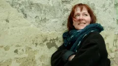 Frau mit braunem Haar lächelt und trägt einen blauen Schal vor einer alten Mauer.