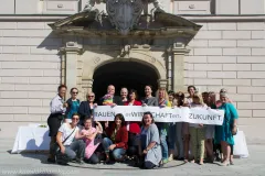 Eine Gruppe lächelnder Menschen hält ein Banner vor einem historischen Gebäude.