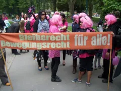 Personengruppe mit rosa Perücken, hält Banner: "Bleiberecht, Bewegungsrecht für alle!".