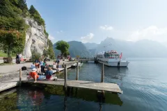 Menschen sitzen auf einem Steg am See; Boot im Wasser, Berge im Hintergrund.