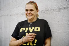 Frau lächelt an Betonwand, trägt schwarzes T-Shirt mit gelbem Text.