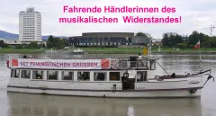 Ein Boot auf einem Fluss mit einem Banner an Deck, städtische Kulisse im Hintergrund.