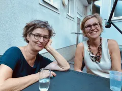 Zwei Frauen sitzen lächelnd an einem Tisch im Freien, neben ihnen Wassergläser.