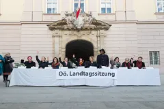 Personengruppe sitzt hinter einem langen Tisch vor einem Gebäude. Einige heben die Hand. Fotografiert.