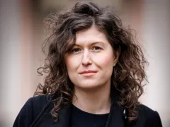 Eine Frau mit lockigem Haar, neutraler Ausdruck, draußen.