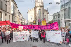 Menschen demonstrieren mit Bannern und Schildern auf einer belebten Straße vor einer Kirche.