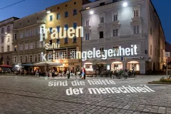 Beleuchtete Fassade mit Projektion auf Kopfsteinpflaster. Abendstimmung, Menschen im Hintergrund.