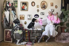 Frau in auffälligem Outfit auf Stuhl, hält Hand eines schwarzen Hundes, viele Fotos an der Wand.
