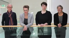 Vier Frauen stehen nebeneinander an einem Geländer und lächeln.