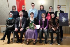 Menschen sitzen und stehen in zwei Reihen, einige halten Blumen und ein Poster.