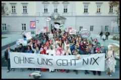 Menschenmenge mit Transparent "OHNE UNS GEHT NIX" vor einem Gebäude, fröhliche Stimmung.