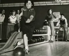 Frau in Bowlingbahn wirft Kugel, beobachtet von vier Personen. Schwarzweiß-Fotografie.
