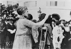 Frau bedient eine alte Kamera, Menschenmenge im Hintergrund, Schwarzweißfotografie.